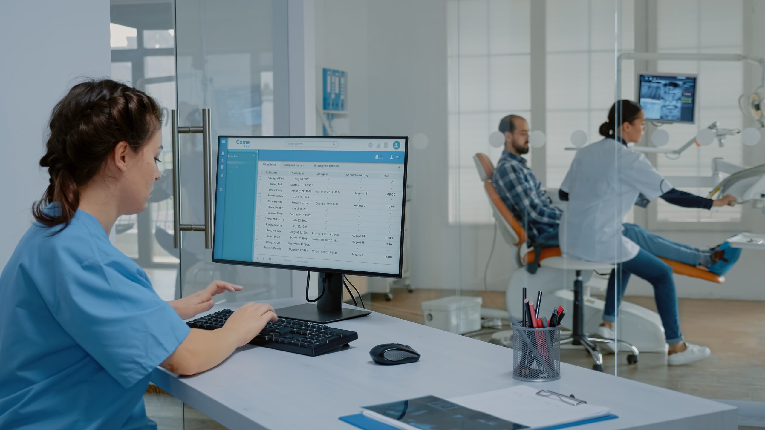 A dental insurance coordinator working through patient records at a front-desk computer while treatment continues in the background